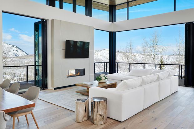 Bright, contemporary double-height living room with expansive black-framed glazing overlooking snow-covered mountains. The room features a large white sectional sofa, pale timber flooring, a white fireplace and media wall, exposed dark structural columns, a glass balustrade, and an adjoining dining area. The open-plan space has a clean, luxurious yet natural feel.