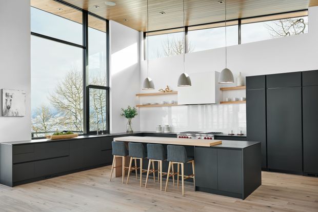 A bright, contemporary kitchen with a large dark island and four upholstered bar stools, floor-to-ceiling corner glazing, white walls, pale timber flooring, and a warm timber-lined ceiling. Matte dark cabinetry runs along the perimeter, with open timber shelves, a white backsplash, and suspended white pendant lights. The glazing provides views of trees and water beyond.
