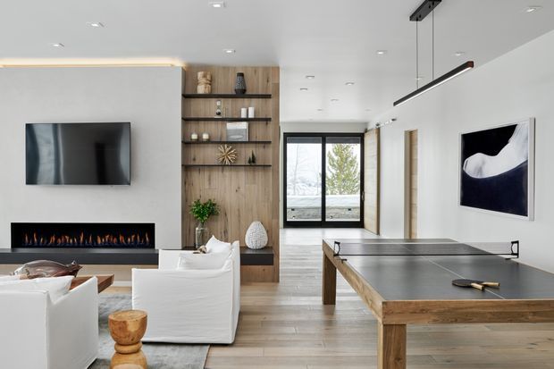 Bright contemporary living room with a wall-mounted television, long linear fireplace, integrated timber shelving and cabinetry, white sofas, and a large timber-framed table-tennis table in the foreground. The room has light wood flooring, white walls and ceiling, recessed downlights, a slim linear pendant, and full-height glazed doors opening toward an exterior view.