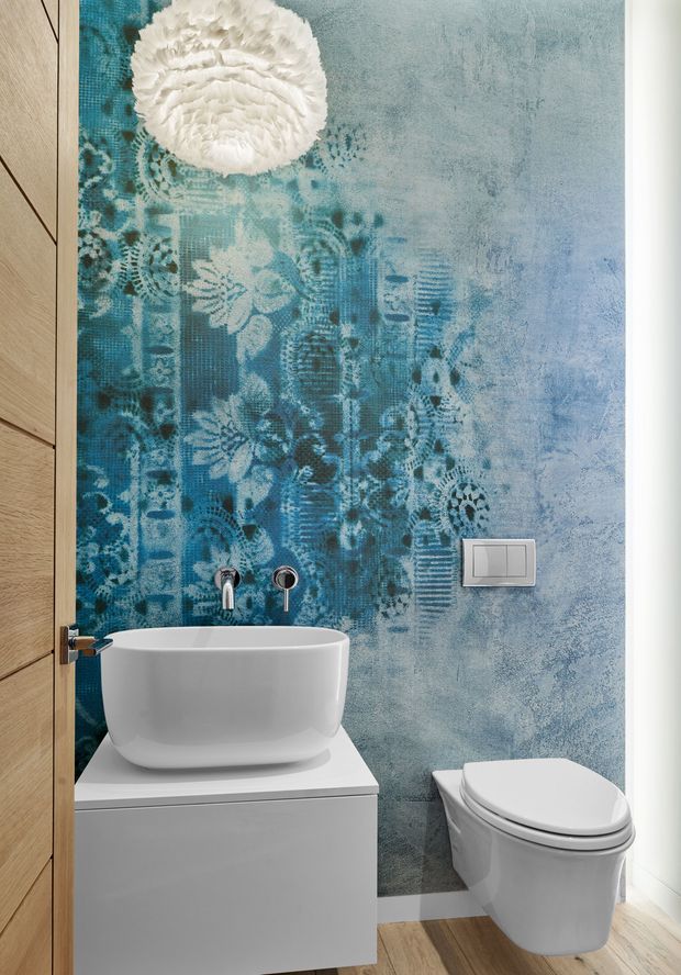 Compact contemporary bathroom featuring a wall-mounted white basin on a minimalist vanity, a white toilet, brushed metal tapware, and a striking blue patterned feature wall with floral and geometric motifs. A timber door and pale timber-look floor add warmth to the otherwise white and blue scheme.