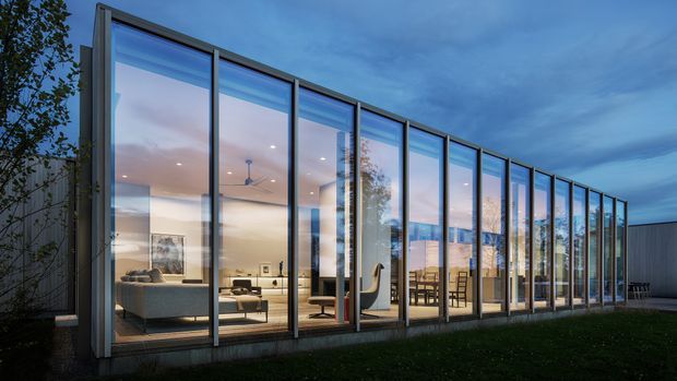 Twilight exterior view of a contemporary, low-profile glass pavilion or extension. The facade is composed almost entirely of floor-to-ceiling glazing in slim dark metal frames, revealing a warmly lit open-plan living and dining area inside. The building sits beside a lawn and garden, with a dark solid-clad volume visible at the right.