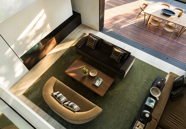 A sunlit contemporary living room viewed from above, arranged around a dark sofa, curved upholstered lounge chair, and central wooden coffee table on a large olive-green rug. Full-height glazed doors open directly to a timber deck with outdoor dining furniture. A minimalist fireplace feature and warm timber flooring add architectural definition.