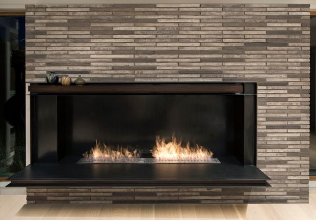 Close-up interior view of a contemporary built-in linear gas fireplace set within a dark surround and wide hearth. The fireplace wall is clad in horizontally stacked, varied-tone brown and grey stone or brick veneer, with a dark timber mantel shelf and pale timber flooring below. Small decorative objects sit on the mantel, and glazing is visible at the left edge.