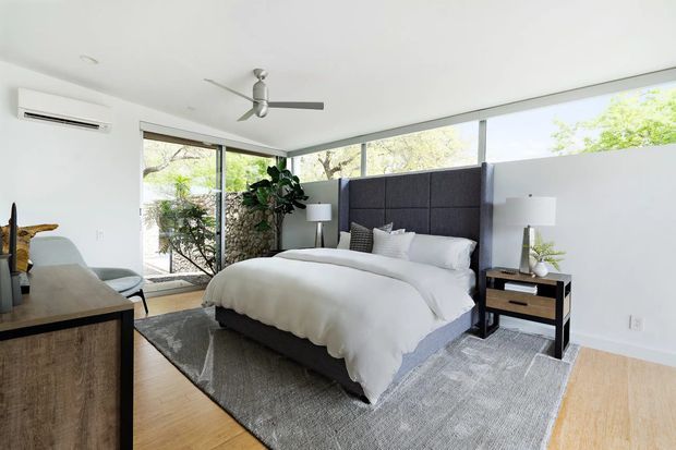 Bright contemporary bedroom with a large upholstered bed and dark charcoal headboard, white bedding, bedside tables and lamps, full-height corner glazing, pale timber flooring, and a large grey area rug. A ceiling fan and split-system air conditioner are visible, with greenery outside the windows.