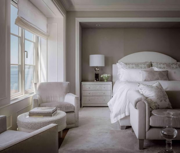 A refined, softly lit bedroom with a large upholstered bed set into a recessed wall alcove, layered white bedding, pale taupe walls, and matching upholstered seating. Tall multi-pane windows with a Roman blind bring in natural light, while a small bedside chest, table lamp, patterned ottoman, and glass side table complete the calm, elegant interior.