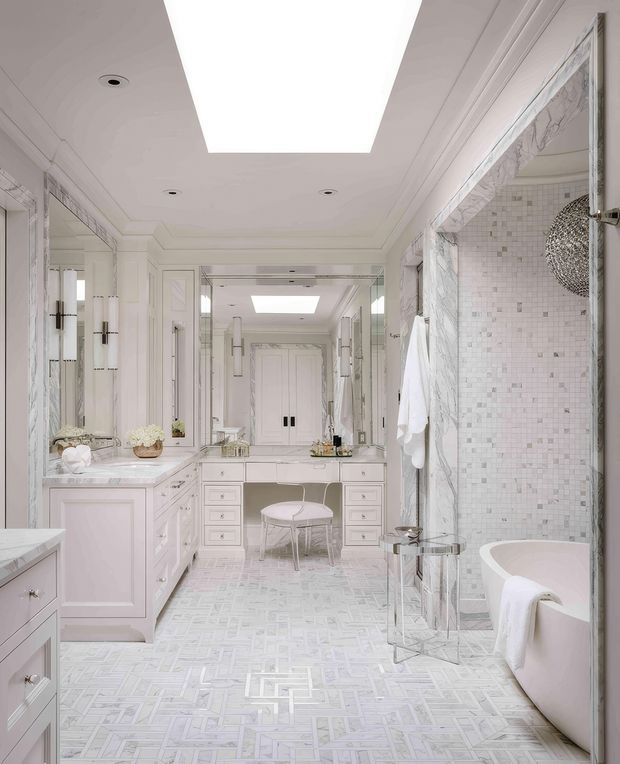 A luxurious, bright bathroom with a symmetrical double vanity, extensive mirrors, pale marble-look wall surrounds, white built-in cabinetry, patterned light stone or tile flooring, a glass-enclosed shower, and a freestanding oval bathtub. Decorative moulded ceiling detailing and a large rectangular skylight add an airy, refined feel.