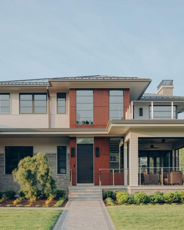 Front exterior view of a contemporary multi-storey residence with a broad low-pitched roof, mixed horizontal siding and stone-textured wall finishes, tall dark-framed windows, red-toned vertical feature panels, a recessed entry, and a covered outdoor sitting area. A paved walkway leads through landscaped front gardens to the house.