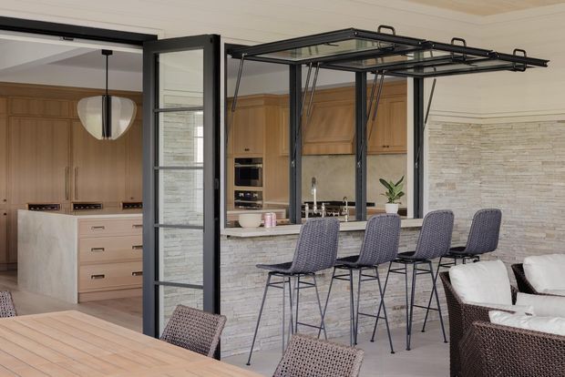 A refined open-plan kitchen and dining area with a bar counter and four woven bar stools. The kitchen features warm timber cabinetry, a pale stone island and counter, integrated appliances, and a light stacked-stone feature wall. Black-framed glazed folding doors separate the space from the dining and living areas, while a contemporary pendant light hangs over the dining table. The palette is warm neutral with contrasting dark metal framing and textured stone surfaces.