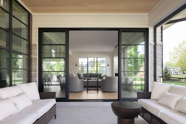 A bright covered outdoor living area with wicker-framed sofas and white cushions, connected through large black-framed glazed sliding doors to a contemporary living room. The space features a pale tiled floor, warm timber-lined ceiling, light horizontal wall cladding, and expansive glazing overlooking a landscaped residential setting.