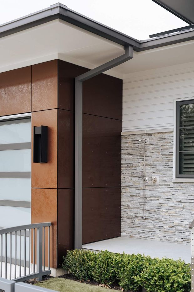 Close-up exterior view of a contemporary residential facade and entry area. The composition features a flat roof with deep white soffits and dark grey metal fascia, rust-toned metal cladding panels around the entry, white horizontal weatherboard siding, a stacked light-grey stone feature wall, a modern black wall light, dark-framed glazing, and a neat hedge beside a metal balustrade.