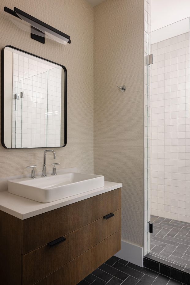 A contemporary bathroom featuring a wall-mounted dark timber vanity with a white rectangular vessel basin and chrome tapware. A rounded-rectangle mirror and slim black wall light sit above the vanity. To the right is a frameless glass walk-in shower with white grid-pattern wall tiles and dark grey herringbone floor tiles. The room has a refined, neutral palette with strong black, white, timber, and grey accents.