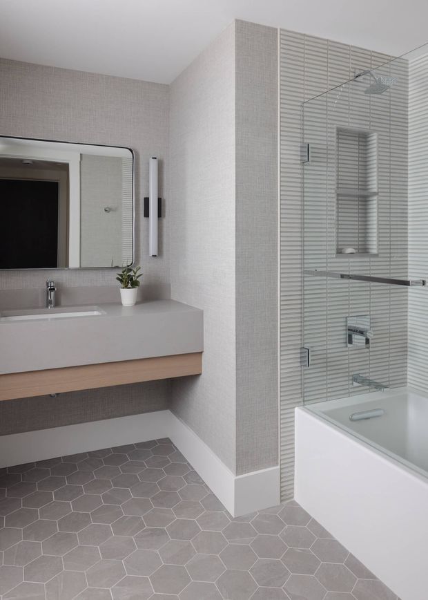 A bright, contemporary bathroom featuring a wall-mounted floating vanity with integrated basin, rounded-corner mirror, chrome tapware, and a small potted plant. The room has grey hexagonal floor tiles, textured pale grey wall finish, and a bathtub with a glass shower screen, recessed shelving, linear wall tiles, and chrome fixtures.