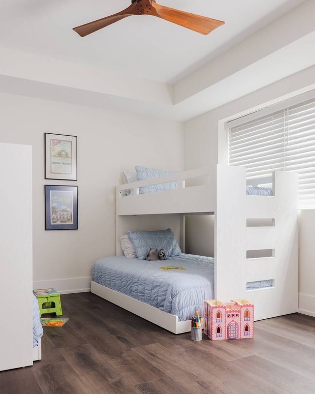 Bright children's bedroom featuring a white built-in bunk bed with blue bedding, pale neutral walls, dark timber-look flooring, framed artwork, white plantation blinds, and a timber ceiling fan. The room has a clean, playful, contemporary feel with generous floor space.