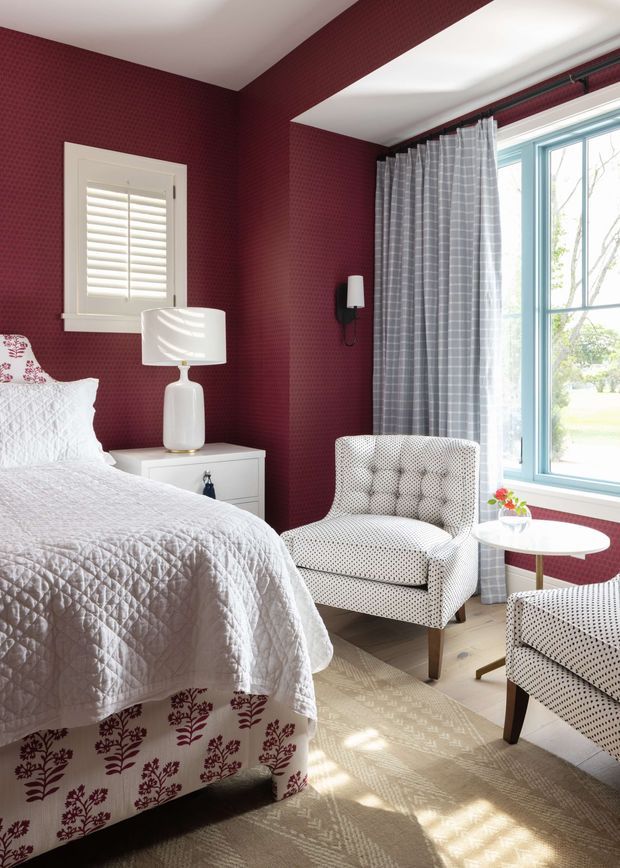 A bright, traditional-style bedroom with deep burgundy patterned walls, white plantation shutters, and large blue-trimmed windows dressed with grey checked curtains. The room contains a neatly made bed with a white quilt and floral bed skirt, a white bedside table and lamp, patterned upholstered armchairs, a small round side table, and a beige patterned rug. Sunlight creates a warm, inviting atmosphere.