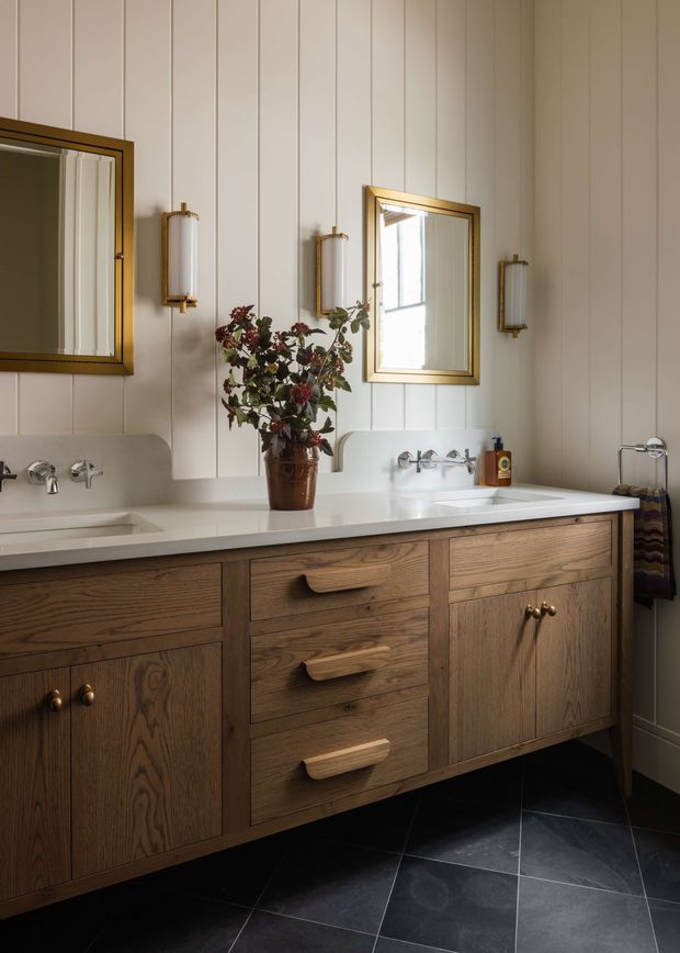 A refined bathroom vanity with a long white benchtop, integrated double basins and wall-mounted taps. The vanity features warm, dark timber cabinetry with sculptural timber handles, set against vertically panelled pale walls. Gold-framed mirrors and matching wall sconces add a traditional decorative character, while large dark floor tiles provide contrast.