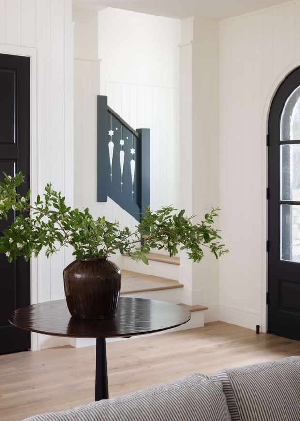 Bright, refined entryway with white vertical wall panelling, pale timber flooring, a black paneled front door and matching arched glazed door, and a short staircase with a dark decorative balustrade. A round dark wood table with a large vase of leafy branches sits in the foreground, with the edge of a striped sofa visible. The space combines crisp white surfaces, black accents, natural timber and organic greenery.