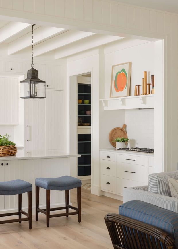 Bright white kitchen and living area with shaker-style cabinetry, vertical wall panelling, exposed painted ceiling beams, a compact cooking alcove, open shelving, and a peninsula with two upholstered stools. A woven armchair and sofa edge are visible in the foreground. The room has pale timber flooring, warm wood accents, and colourful artwork.