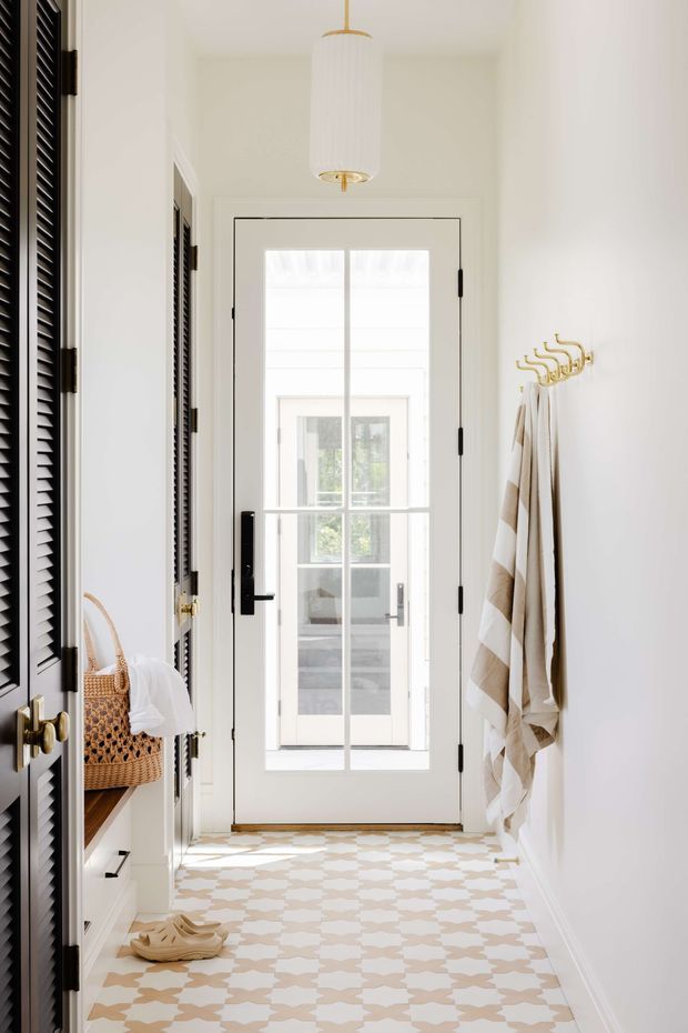 Bright, narrow residential hallway or entry passage with white walls, a glazed exterior door at the end, dark louvred built-in cabinetry on the left, and brass hardware. Patterned beige-and-white floor tiles, a white pendant light, woven basket storage, and wall-mounted brass hooks with hanging towels create a clean, refined look with strong natural light.