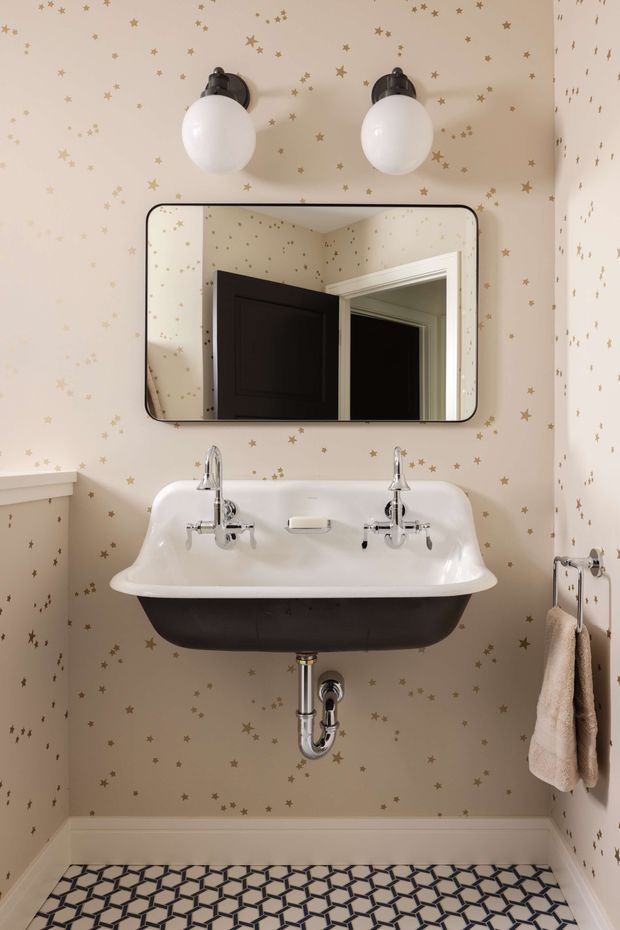 A compact, charming bathroom centered on a wall-mounted double basin with chrome taps and exposed plumbing. The walls are covered in warm beige wallpaper scattered with small metallic gold stars. Above the basin is a rounded rectangular mirror with a dark frame, flanked by two globe wall lights. The room has white trim and a bold geometric black, white, and grey patterned floor, with a small towel rail on the right.
