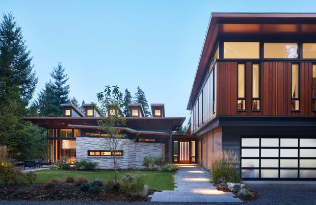 Exterior view of a contemporary multi-wing residence set within landscaped grounds. The home features strong horizontal and vertical volumes, warm timber cladding, dark metal detailing, expansive glazing, clerestory-like windows, and a prominent integrated garage. A lower connecting wing has a low-pitched roof with multiple raised dormer or clerestory elements, while stone walling and illuminated paths define the entry landscape.