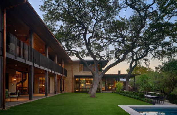 A large two-storey residential building arranged around a landscaped courtyard and swimming pool. The architecture features extensive warm timber cladding and exposed timber soffits, dark slender balcony railings and posts, broad glazed openings, and a low contemporary wing extending into the garden. Mature trees dominate the courtyard, creating a sheltered, tranquil outdoor setting.