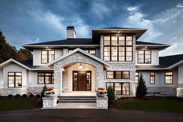A large two-storey residential home with a symmetrical, grand façade. The exterior combines white horizontal weatherboard cladding with light grey stone masonry, including a prominent arched entry porch, stone chimney, dark low-pitched roof, and extensive black-framed glazing. Formal landscaping and a broad paved approach frame the entrance beneath a dramatic cloudy sky.