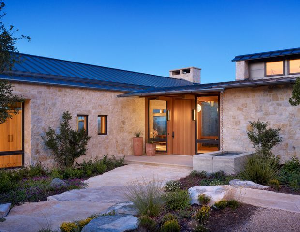 Single-storey stone-clad residence with a low-pitched dark metal roof, warm timber entry door and glazed sidelights, and a small raised upper-level section at the right. The entry is approached by a pale stone-paved path through a landscaped garden with native-style planting and boulders. The exterior is photographed at dusk against a clear blue sky.