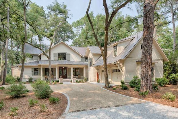 Exterior view of a large light-coloured weatherboard home set among mature trees, with multiple pitched gabled roof forms, dormer-like upper-level windows, a covered front porch, and an attached garage. A curved paved driveway and landscaped garden beds lead toward the house.