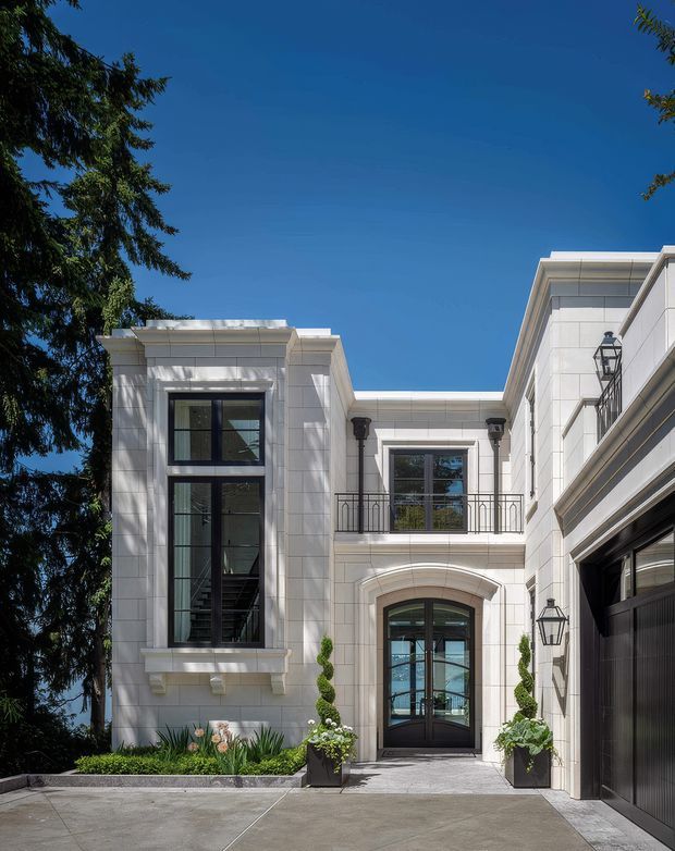 A formal white stone-clad residential facade viewed from the driveway courtyard. The multi-level entry features tall black-framed windows, a glazed arched front door, wrought-style balcony railings, classical trim and pilasters, decorative wall lanterns, and an integrated dark garage door. Sculpted topiary and garden planting soften the paved approach, with a large evergreen tree framing the left side.