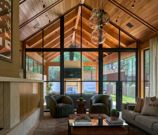 Warm, timber-lined living room with a dramatic pitched ceiling and exposed rafters, framed by extensive black steel or aluminium glazing. The room includes a fireplace with timber cabinetry, two deep green lounge chairs, a neutral sofa, patterned rug, and a central glass coffee table. Glazing opens visually to a leafy enclosed outdoor area, while sculptural glass pendant lights hang from the ceiling.