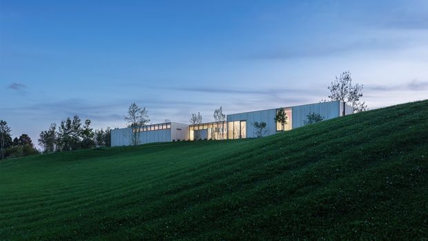 A long, low modern building is partially embedded along a gently sloping grassy site. The facade features pale blue-grey vertical cladding, extensive warm-lit glazing, and a simple flat or very low-pitched roofline. Sparse young trees frame the building, shown at dusk beneath a blue sky.
