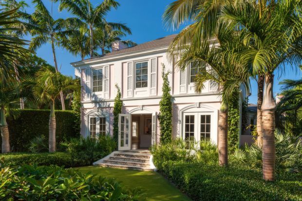 A two-storey pale rendered residence with a symmetrical classical facade, white-trimmed multi-pane windows, shutters, arched ground-floor openings and a central entrance. The home is surrounded by manicured tropical landscaping, including palms, clipped hedges and dense planting, under a clear blue sky.