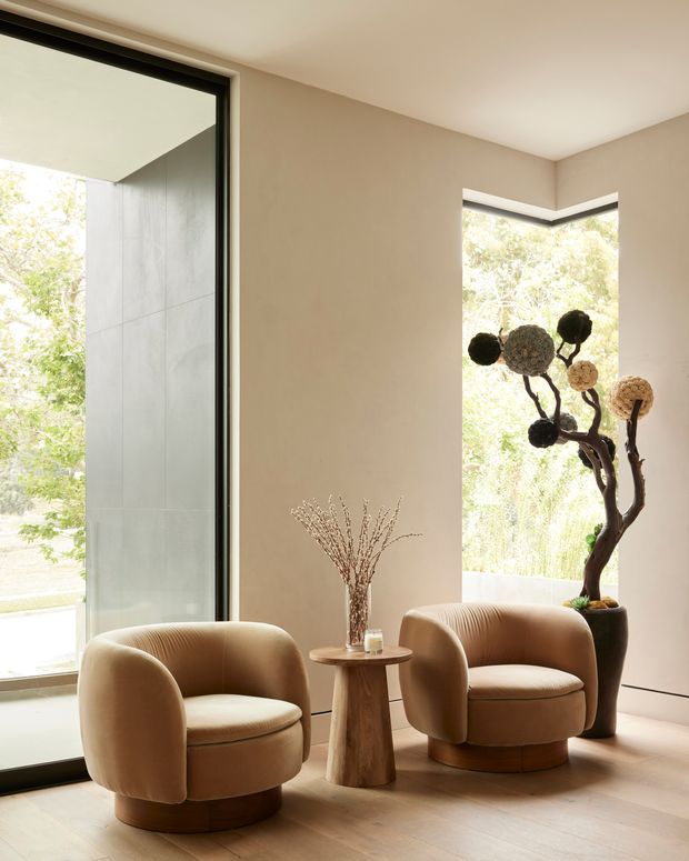 A softly lit minimalist sitting area with two rounded upholstered lounge chairs in warm taupe, arranged around a small sculptural wooden side table. Floor-to-ceiling black-framed glazing and a corner window bring in abundant natural light and views of greenery. A tall sculptural indoor plant and simple dried-branch arrangement add organic accents to the refined neutral interior.