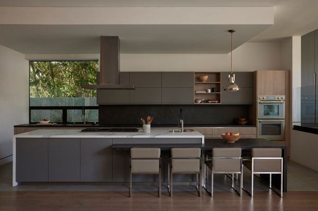 Contemporary open-plan kitchen with a large dark island, integrated cabinetry, stone benchtops, a black splashback, timber-toned flooring, built-in stainless-steel appliances, and four bar stools. A broad window brings in views of greenery, while a pendant light and recessed ceiling detail add understated architectural character.