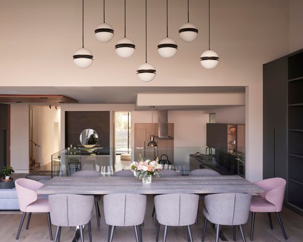 A contemporary open-plan dining and kitchen interior viewed from the living area. A long stone dining table is surrounded by upholstered dining chairs in pale grey and blush pink, with a sculptural cluster of globe pendant lights overhead. Behind the dining area is a dark-toned kitchen with island, cabinetry, integrated appliances and a glass partition, while dark built-in shelving occupies the right side. The room has a warm, refined palette and generous ceiling height.