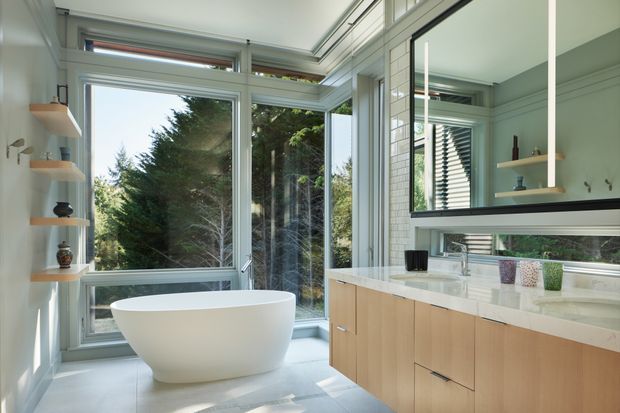 Bright contemporary bathroom with a freestanding white soaking tub positioned beside expansive corner windows overlooking trees. A floating light-wood vanity with a pale stone benchtop spans the right side, beneath a large mirror and horizontal window. Open shelving with small decorative objects lines the left wall, while white tiled walls, pale flooring, and clerestory glazing create an airy, nature-connected setting.