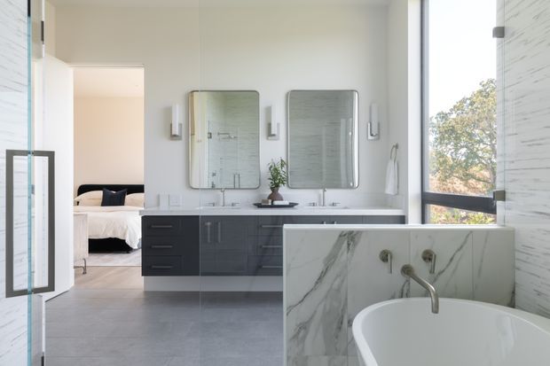 A bright, spacious contemporary bathroom with a freestanding white bathtub in the foreground, a double vanity with dark built-in cabinetry and two rounded rectangular mirrors, wall-mounted lights, and large-format grey flooring. A bedroom is clearly visible through the open doorway, while tall glazing brings in natural light and views of trees. The bathroom features a marble-look tiled wall and bathtub surround with black and grey veining.