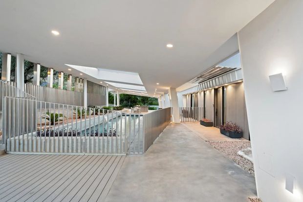 A covered contemporary exterior walkway or courtyard passage with a smooth concrete floor, pale vertical metal balustrades, timber decking, integrated garden beds, and a linear water feature or pool visible to the left. White rendered walls, recessed ceiling lights, narrow clerestory-like glazing, and vertical screening create a clean, sheltered architectural circulation space.