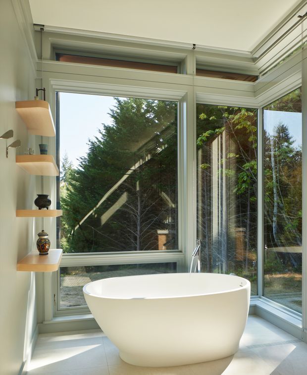 Bright contemporary bathroom with a freestanding white oval bathtub positioned in a glazed corner. Large windows and glass doors provide extensive views of surrounding trees, while pale green-grey window frames, light tiled flooring, and built-in curved timber display shelves create a calm, nature-connected setting.