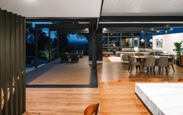 Open-plan contemporary interior showing a kitchen island in the foreground, a dining area and living room beyond, and expansive dark-framed glazing opening onto a timber outdoor deck. Warm timber flooring and cabinetry contrast with white surfaces, dark structural framing, and evening views outside.