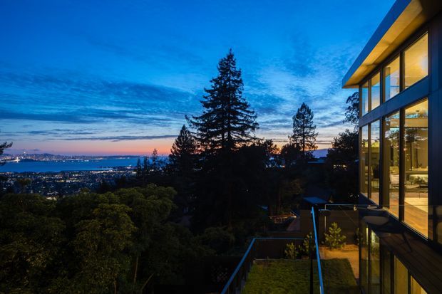 Twilight exterior view of a contemporary multi-level home on a vegetated, elevated site. The building features extensive floor-to-ceiling glazing, dark structural framing, warm illuminated interiors, projecting roof eaves, and a glass-balustraded terrace. A broad coastal or bayside urban panorama is visible beyond the trees.