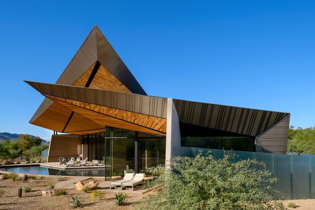 A striking contemporary desert residence with sharply folded, angular roof planes and dramatic cantilevered eaves. The facade combines warm timber soffits and cladding with dark metal roof surfaces, extensive glazing, and a partially screened outdoor living area. A swimming pool, patio furniture, gravel landscaping, and drought-tolerant planting are visible in the foreground, with mountainous terrain in the distance.