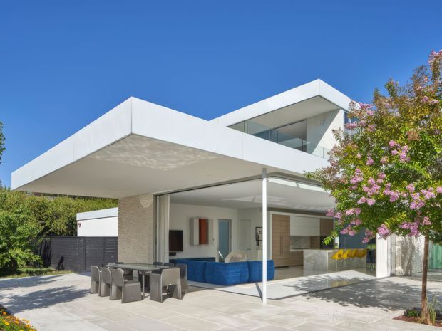 Exterior view of a contemporary two-storey flat-roof house with strong horizontal cantilevered roof planes, extensive glazing, and an open ground-floor entertaining area. The pale rendered facade and white roof edges frame a covered patio with outdoor dining and seating, while a bright blue sofa and warm timber-toned interior joinery are visible through the open glazing. A flowering tree borders the right side of the property.