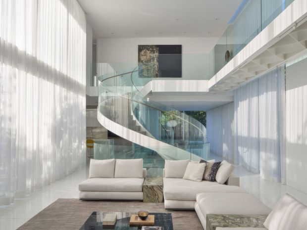 A bright, spacious contemporary living room with a large cream sectional sofa, glass coffee table, sheer floor-to-ceiling curtains, and pale stone or polished flooring. A dramatic curved staircase with a glass balustrade rises through a double-height void, with a mezzanine walkway and extensive glazing visible beyond. The palette is predominantly white, cream, pale grey, and transparent glass, creating an airy and refined atmosphere.