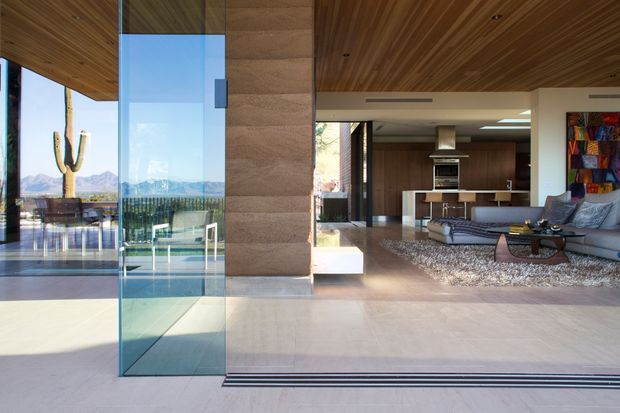 Wide view from an open glazed threshold into a contemporary open-plan living and kitchen area. The room features a warm timber-lined ceiling, large floor-to-ceiling glazing, pale stone or concrete-like flooring, a stone-clad structural column, streamlined dark cabinetry, a sectional sofa, and a textured rug. A terrace and desert landscape with a cactus are visible beyond the glazing.
