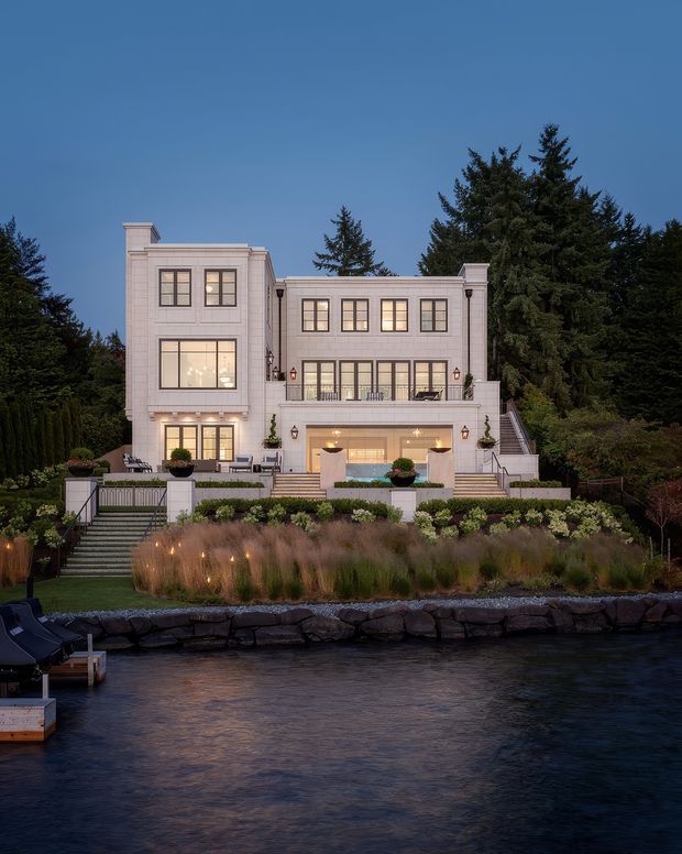 A large, symmetrical waterfront residence viewed from across the water at dusk. The pale multi-level facade features classical detailing, tall gridded windows, balconies, terraces, broad staircases, and warm architectural lighting. Formal landscaped gardens, stone shoreline edging, and a small dock frame the foreground, with dense evergreen trees behind the house.