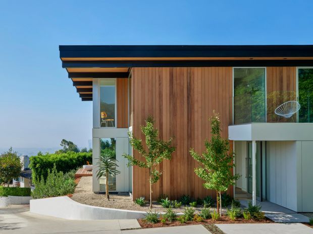Exterior view of a contemporary two-storey residence with a projecting flat roof, exposed dark roof edges and warm timber soffit detailing. The facade combines vertical timber cladding with light rendered or panelled sections and large glazed openings. A landscaped front garden, curved low retaining wall and paved approach are visible, with a distant ocean or blue horizon beyond the property.