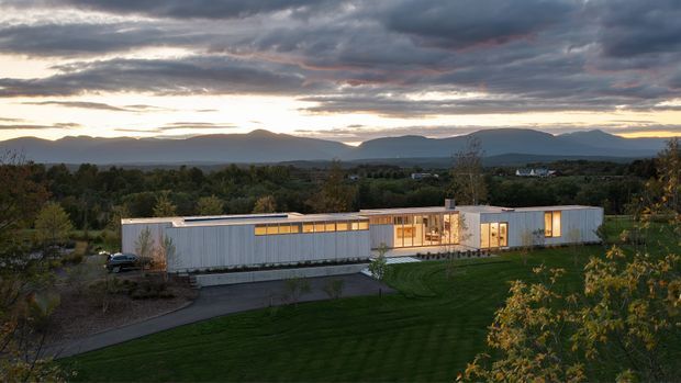 A long, low modern residence set within a landscaped suburban or rural site, photographed at dusk. The building has a strongly horizontal composition, pale ribbed metal-like cladding, a low flat roof, extensive warm-lit glazing, and a central glazed entrance or living zone. A driveway and broad lawn occupy the foreground, with trees and distant mountains beneath dramatic cloud cover.