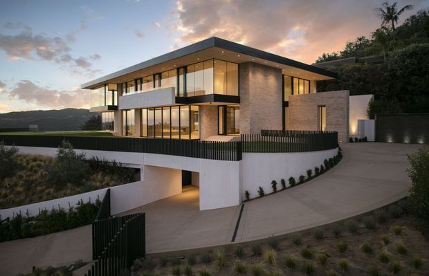 A contemporary multi-level modern residence set on a sloping site, photographed at dusk. The house features a broad flat roof with deep overhangs, extensive floor-to-ceiling glazing, pale masonry feature walls, dark metal window frames and vertical fencing, plus a partially enclosed lower-level garage or undercroft. Warm interior lighting contrasts with the cool evening exterior and surrounding landscaped hillside.