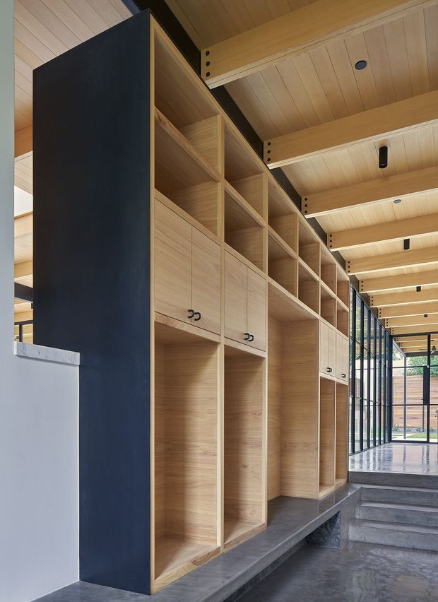 A modern corridor or entry zone lined with bespoke light timber built-in cabinetry and open shelving. The joinery incorporates tall storage bays, cupboards with dark handles, and integrated bench-like sections. Exposed timber ceiling boards and substantial timber beams continue overhead, with black steel structural and glazed framing along the right side. Polished grey concrete flooring and steps lead toward a bright glazed outdoor area.